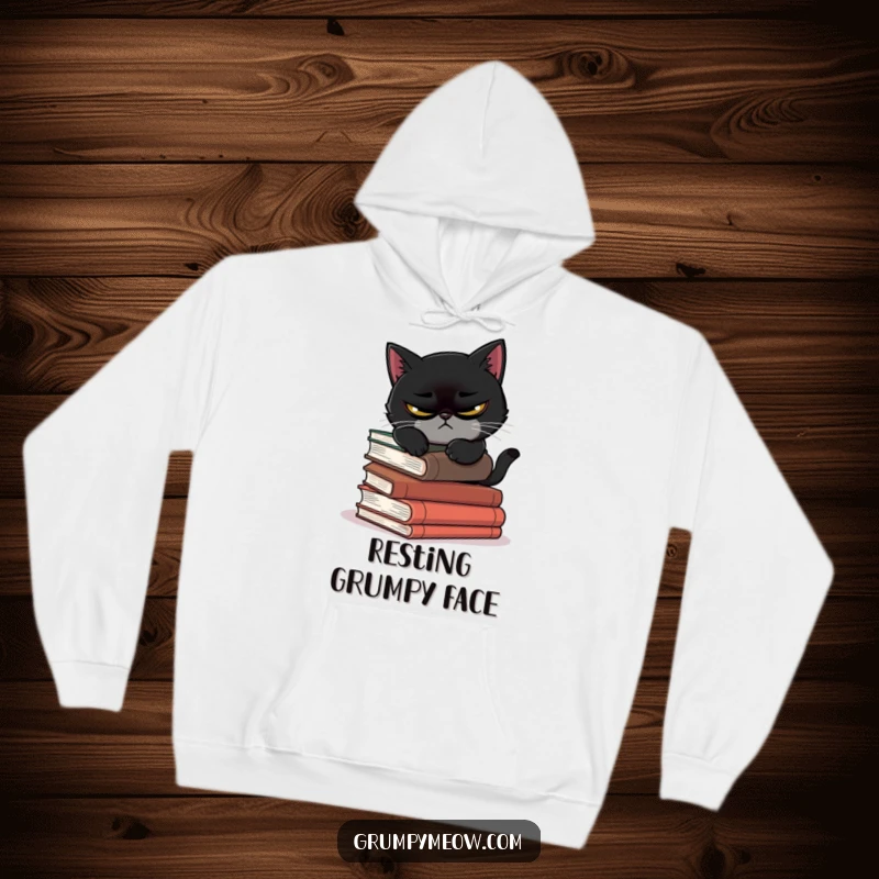 Funny hoodie featuring a scowling black cat attempting to nap precariously on a pile of books, ideal for cozy humor.